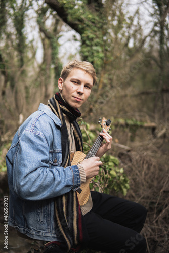An adult smiling man plays the ukulele in the woods by the river. A strong guy sits and plays a small guitar, against the background of the forest. A happy person plays a musical instrument.