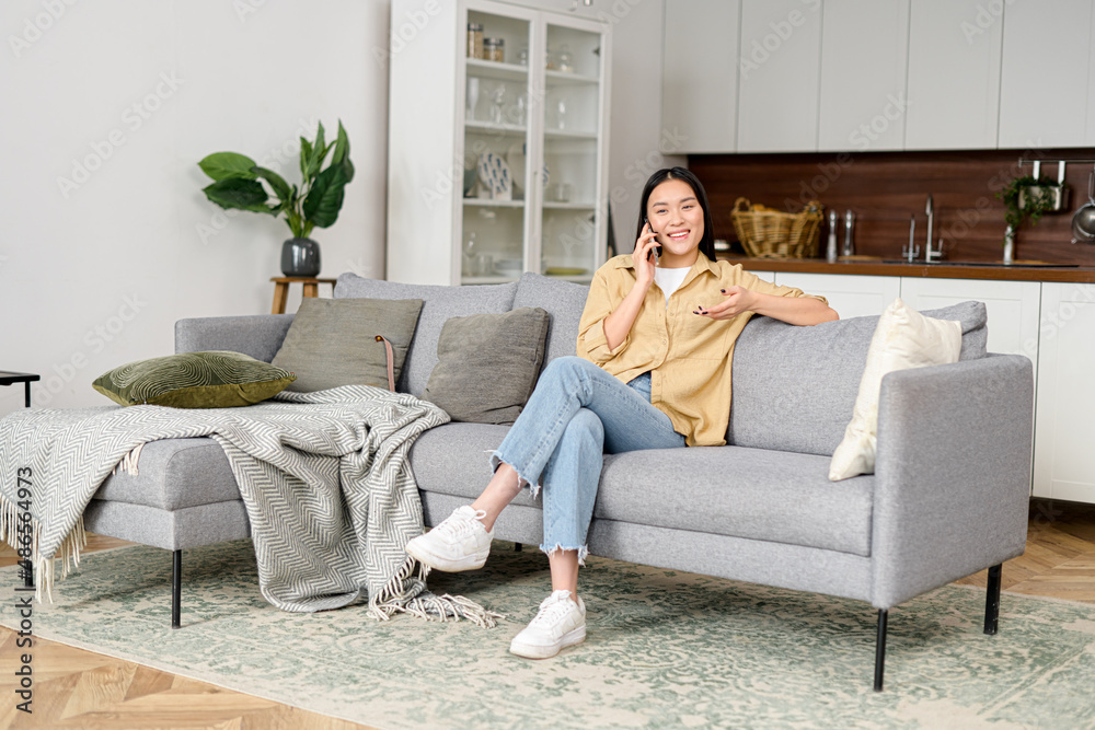 Charming asian woman talking on the smartphone sitting on the cozy couch at home. Smiling young woman chatting with friend on mobile phone, has pleasure phone conversation Charming asian woman talking on the smartphone sitting on the cozy couch at home. Smiling young woman chatting with friend on mobile phone, has pleasure phone conversation