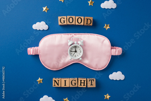 Top view photo of one big pink blindfold and small white alarm on it with scattered confetti in shape of stars and clouds and wooden cubes with words on the deep blue background photo