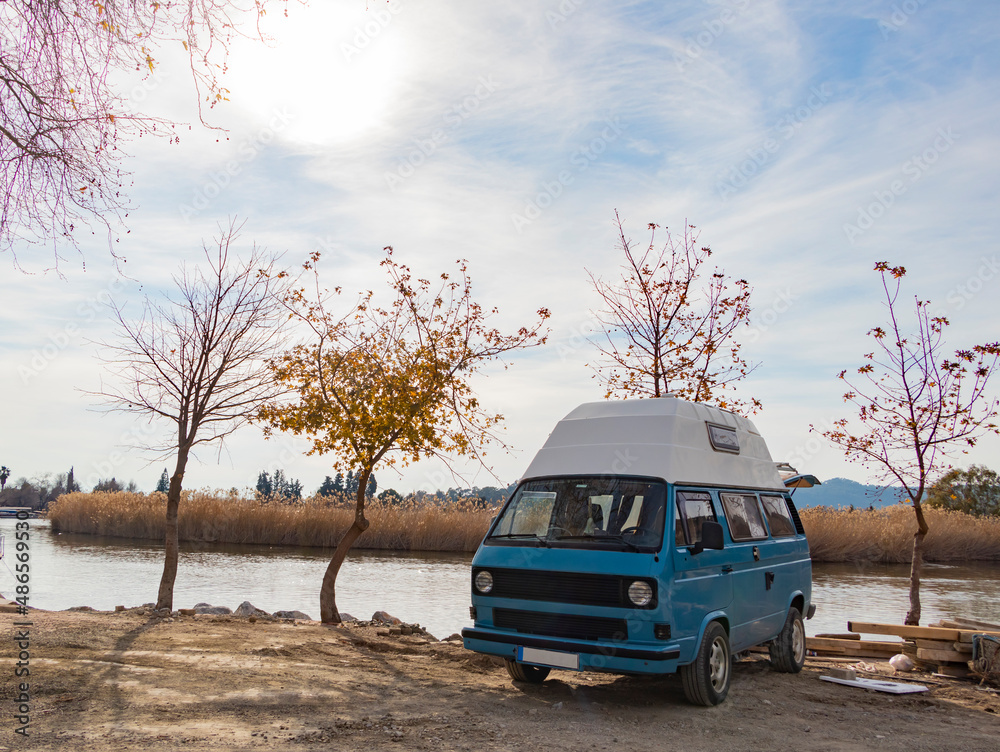 Sweet blue caravan parked by the lake. Autumn season. Naked tree. Beautiful landscape.