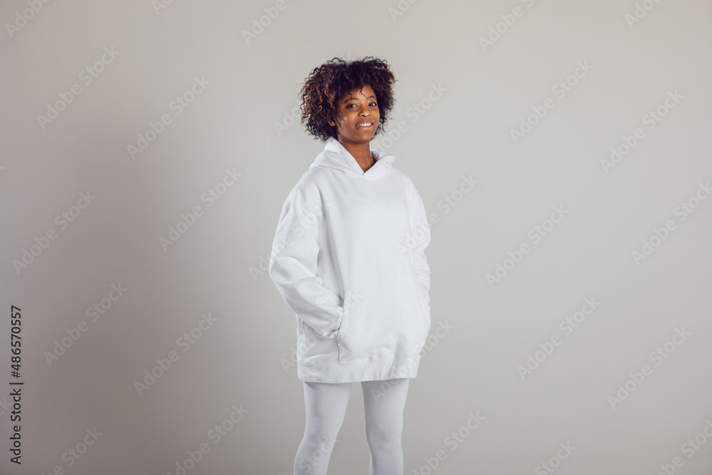 African American woman with puffy hair in a white hoodie and leggings stands on a white background in a photo studio. African American woman with puffy hair in a white hoodie and leggings stands on a white background in a photo studio.