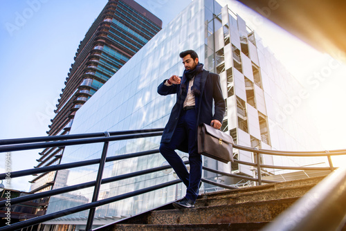 Attractive young businessman in stylish suit with briefcase in business district walking down the stairs while looking at his watch