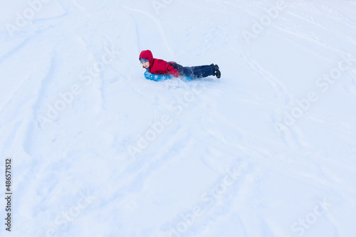 Boy sliding on blue tubing on stomach in winter clothes among white snow, distant view, copyspace. Inflatable sleigh rides.