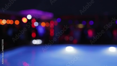 Blurred lights of bright garland hanging on building near swimming pool. Party with neon decorations at night. Calm water ripple on pool surface