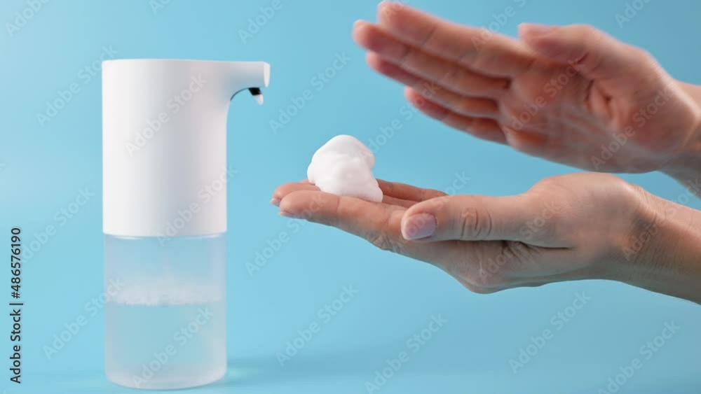Hands of young woman touching soap suds on blue background. Concept of hand washing, hygiene, health, virus and bacteria protect. White foam soap, automatic soap dispenser Hands of young woman touching soap suds on blue background. Concept of hand washing, hygiene, health, virus and bacteria protect. White foam soap, automatic soap dispenser