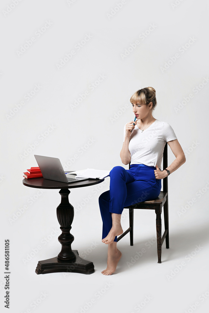 Thoughtful young female student sitting at desk working or studying remotely online looking on laptop screen with interest biting pen and thinking. Distant learning or remote work from home.