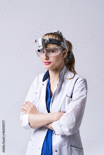Confident ENT doctor wearing surgical headlight head light and protective glasses. Portrait of female otolaryngologist or head and neck surgeon standing arms crossed on light grey background.