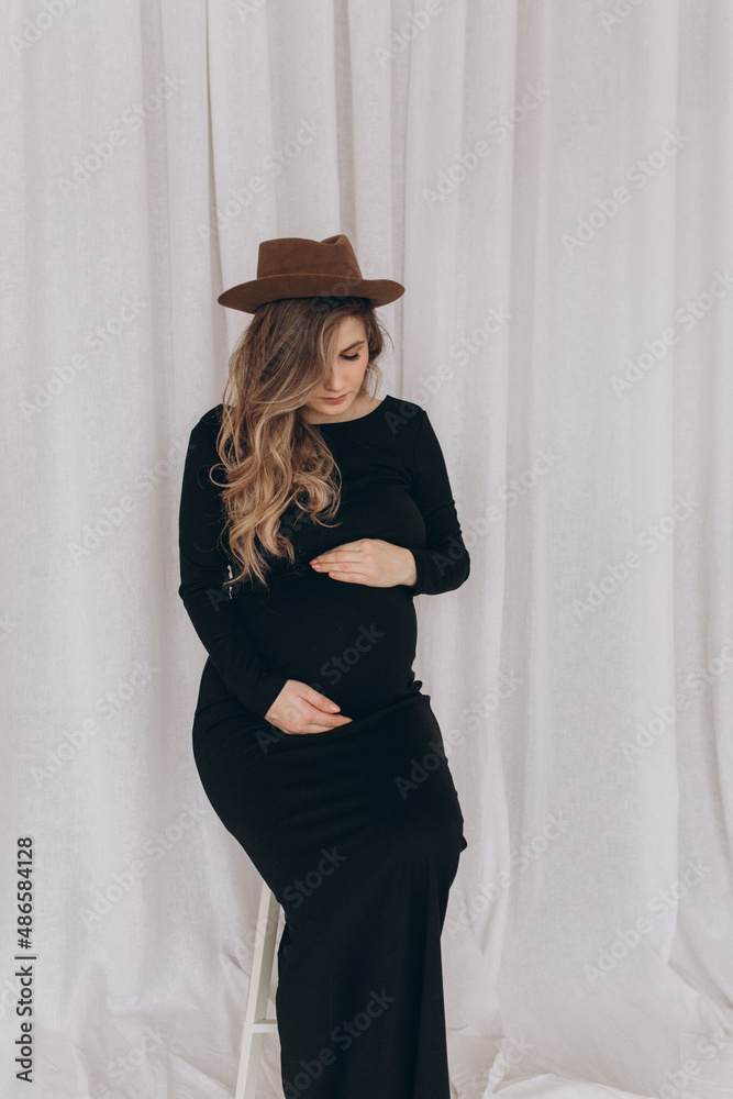 Pregnant woman in black dress sitting on the chair in the studio with white background. Pregnant woman touching belly. Pregnant woman in black dress sitting on the chair in the studio with white background. Pregnant woman touching belly.