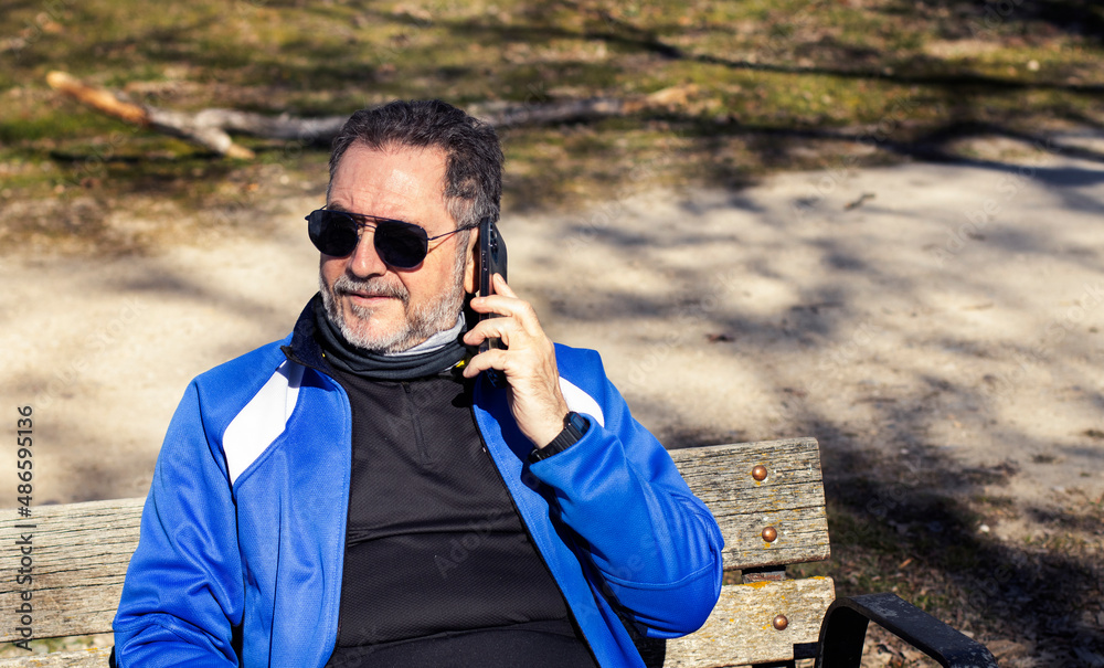 middle-aged man with sunglasses and a white beard, making a phone call with his smart phone sitting on a park bench middle-aged man with sunglasses and a white beard, making a phone call with his smart phone sitting on a park bench