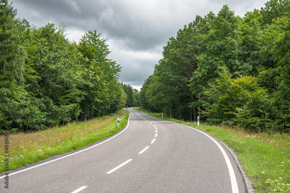 Fototapeta premium Deserted winding road through a forest under storm clouds in the countryside of Germany in summer