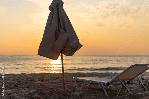 Evening on the beach. A closed beach umbrella obscures the setting sun. A chaise lounge on the sandy beach in the evening