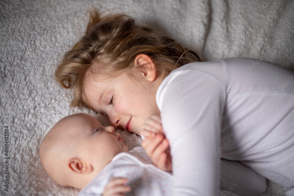 Happy children, toddler and older sister, hugging at home on a white blanket, smiling, shot from above. blonde girl hugging her newborn sister Happy children, toddler and older sister, hugging at home on a white blanket, smiling, shot from above. blonde girl hugging her newborn sister