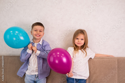 The boy smiles, the girl looks at the camera. A young boy in a blue shirt and a girl in a white T-shirt and blue jeans are standing on the sofa holding balloons