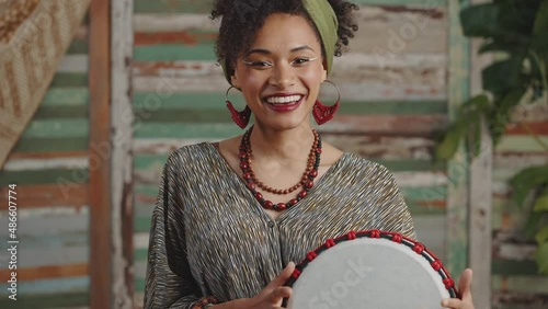 Ethnic traditions. Young happy stylish african american woman drumming on djembe with fingers and smiling to camera photo