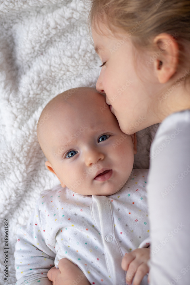Happy children, toddler and older sister, hugging at home on a white blanket, smiling, shot from above. blonde girl hugging her newborn sister Happy children, toddler and older sister, hugging at home on a white blanket, smiling, shot from above. blonde girl hugging her newborn sister