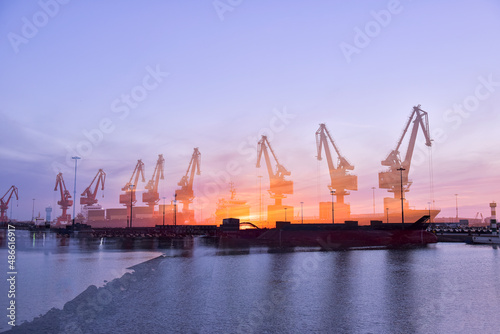 In the setting sun, the crane at the coastal wharf is lifting goods for the cargo ship, with double photographic effects.