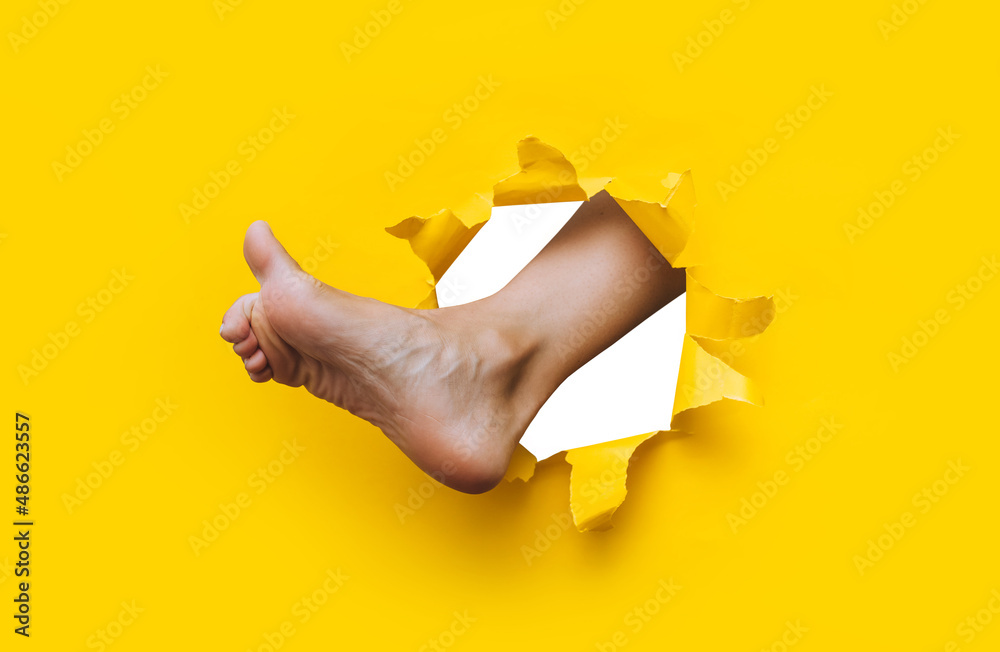 A right woman's foot appears in a torn hole in yellow paper and tries ...