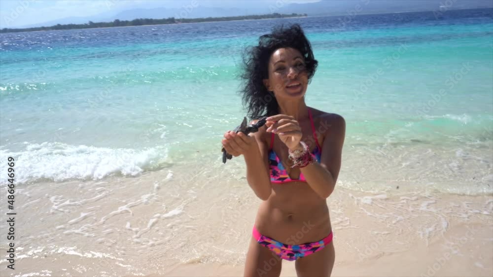 Cheerful woman holding small turtle in hands, showing to camera cute ocean creature, waves on the tropical beach