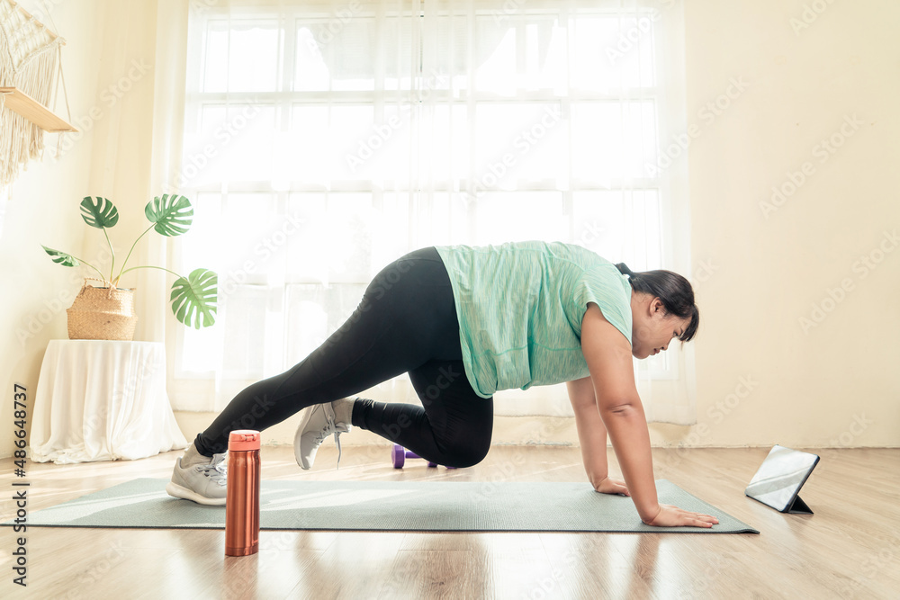 Asian young female plus size in sport wear effort doing exercise on yoga mat in living room. Watching training in loss weight course online video in tablet. Healthy lifestyle concept.