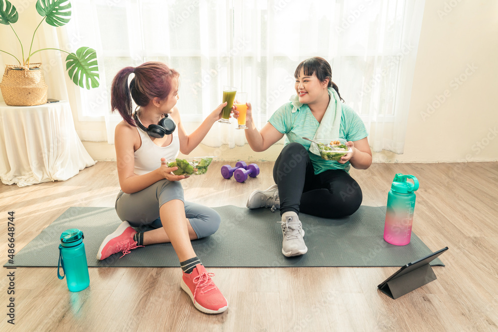 Dieting and exercise at home concept. Two Asian women body size different in sport wear sitting healthy eating vegetable salad and drinking fruit juice after exercise at home together. Dieting and exercise at home concept. Two Asian women body size different in sport wear sitting healthy eating vegetable salad and drinking fruit juice after exercise at home together.