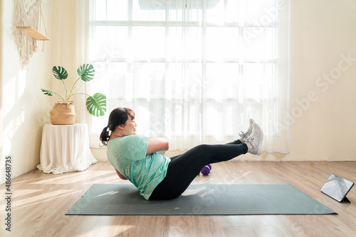 Asian young female plus size in sport wear effort exercise training abdominal muscle on yoga mat in living room. Training in loss weight course online video in tablet. Healthy lifestyle concept.