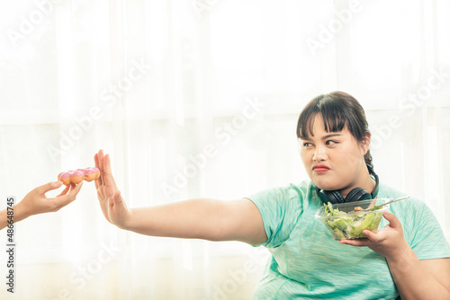 Asian plus size women dieting for the wellness during hand pushing out donut and choosing vegetable salad food for good health. Plus size women and health care concept. photo