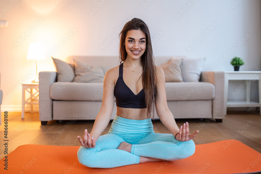 Young attractive smiling woman practicing yoga, sitting in Half Lotus exercise, Ardha Padmasana pose, working out, wearing sportswear, meditation session, indoor full length, home interior Young attractive smiling woman practicing yoga, sitting in Half Lotus exercise, Ardha Padmasana pose, working out, wearing sportswear, meditation session, indoor full length, home interior
