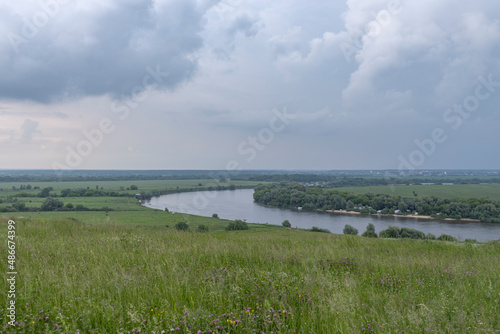 Summer landscape with meadow grass in the foreground. Dark sky with clouds before rain. Bright green grass. Wide river in the background.