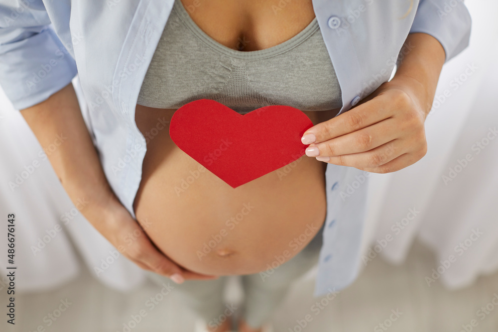 Baby expectation. Young pregnant woman expecting birth of son or daughter holds red paper heart near her belly. Close up of red heart over pregnant tummy in hands of unknown woman. Pregnancy concept. Baby expectation. Young pregnant woman expecting birth of son or daughter holds red paper heart near her belly. Close up of red heart over pregnant tummy in hands of unknown woman. Pregnancy concept.
