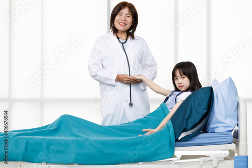 Portrait studio shot of Asian professional successful experienced senior female doctor in white lab coat with stethoscope smiling look at camera visiting little girl patient laying down on clinic bed