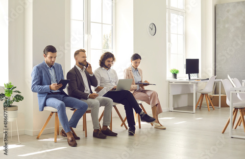 Group of people waiting for job interview or business meeting. Busy men and women sitting in line in office interior, holding CVs and resumes, reading documents, using devices, talking on smartphones