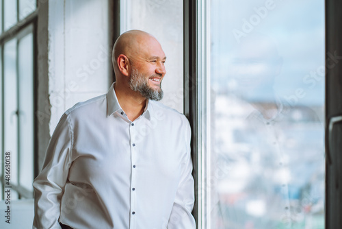 Happy bald bearded mature businessman in white shirt standing near panoramic window and enjoying city view. Successful smiling middle-aged male CEO executive in office, selective focus