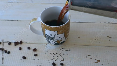 From the coffee maker, black coffee is poured into a cup, scattered coffee beans next to it. Slow motion video.
