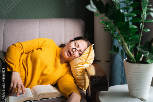 Middle-aged woman in glasses fell asleep while reading book at home, female sleeping with open mouth on sofa near coffee table with houseplant in living room. Sleep deprivation
