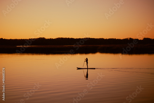 Silhouette of woman doing sup boarding alone on lake at sunset time holding oar in hands with charming yellow orange sky, water and trees in background. Active lifestyle.