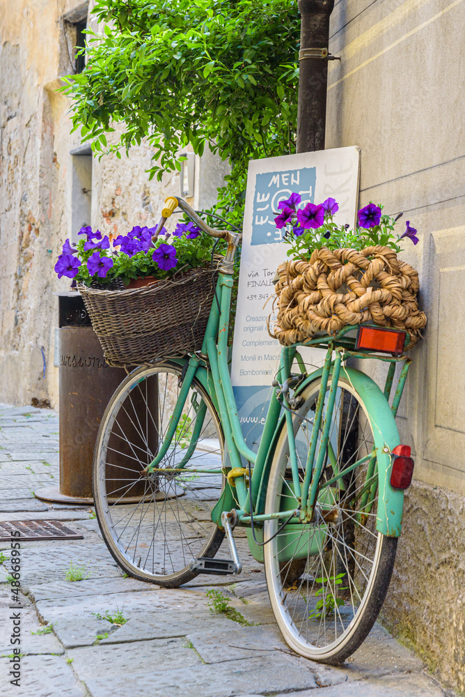 Finalborgo, Finale Ligure, Italy. May 5, 2021. In a small street in the center an old green bicycle leaning against a wall with wicker baskets with flower pots inside.