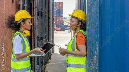 Two African female sisters in safety uniform and hardhat use computer to have a conversation next to a blue shipping container at a cargo storage. Female supervisor provides instruction to trainee