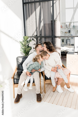 Happy stylish parents sitting on chairs with beautiful kids, spending weekends at home. Loving husband kissing lovely wife, enjoying moments together, happy family concept