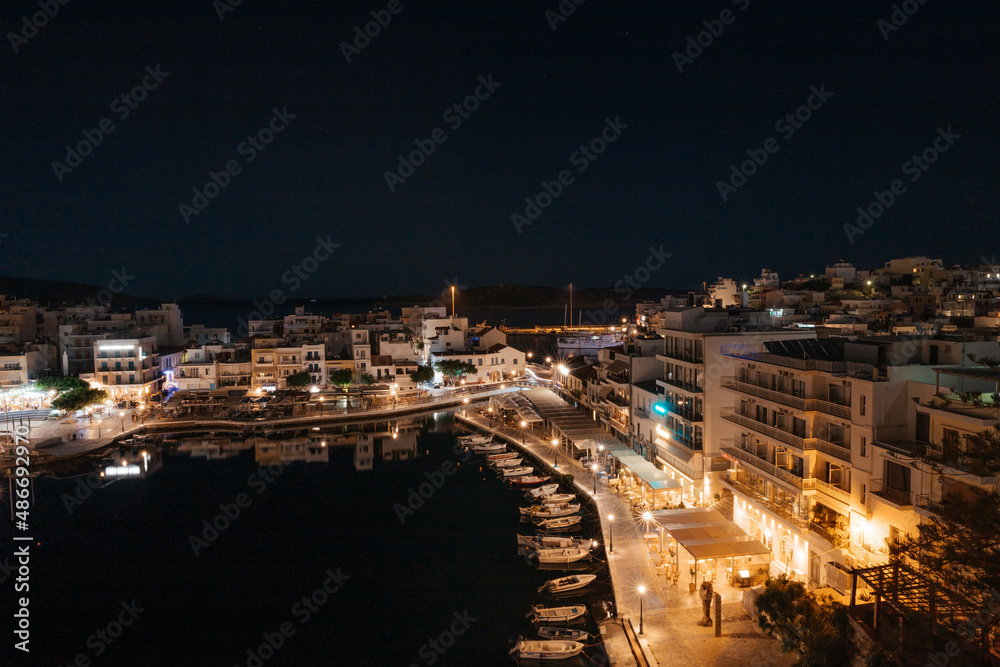 Beautiful night view of small town Agios Nikolaos and Voulismeni lake in Crete island, Greece. Place with waterfront with cafe and restaurants, boats and mountains on the background..