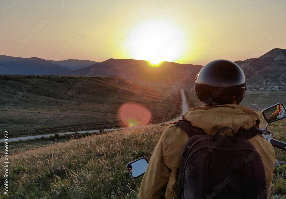 Default. Sunny sunset in the mountains. A man with a backpack sits on a scooter against the backdrop of a mountain range in sun glare Default. Sunny sunset in the mountains. A man with a backpack sits on a scooter against the backdrop of a mountain range in sun glare