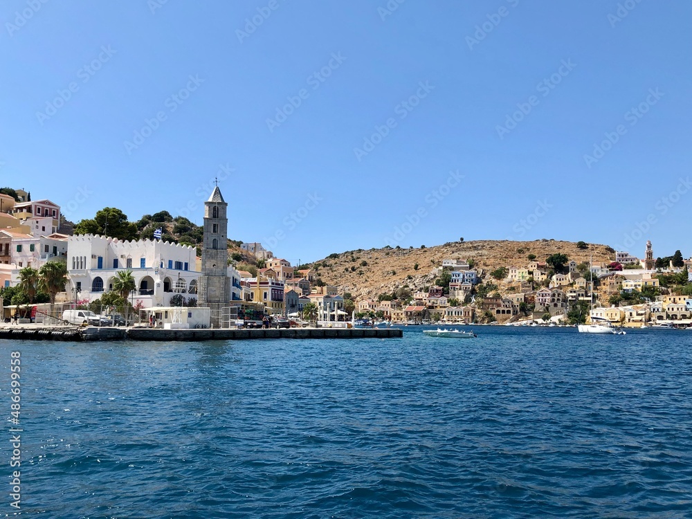 Symi clock tower. Symi island town centre and Aegean sea view. One of small Greek Dodecanese islands. Small marina with boats and yachts. Aegean Sea and island view. Turquoise blue water, colourful bu