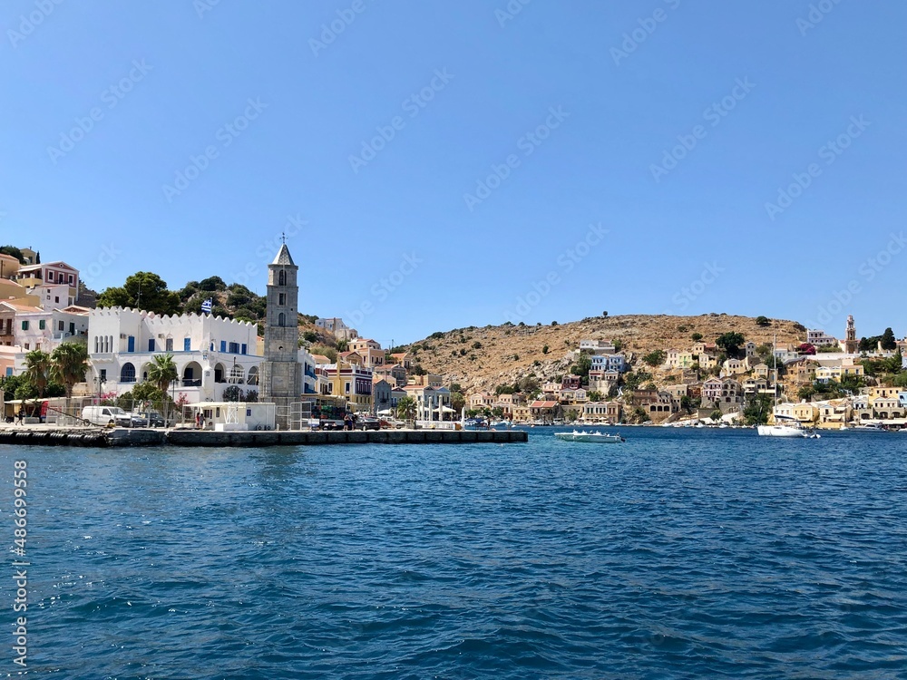 Symi clock tower. Symi island town centre and Aegean sea view. One of small Greek Dodecanese islands. Small marina with boats and yachts. Aegean Sea and island view. Turquoise blue water, colourful bu Symi clock tower. Symi island town centre and Aegean sea view. One of small Greek Dodecanese islands. Small marina with boats and yachts. Aegean Sea and island view. Turquoise blue water, colourful bu