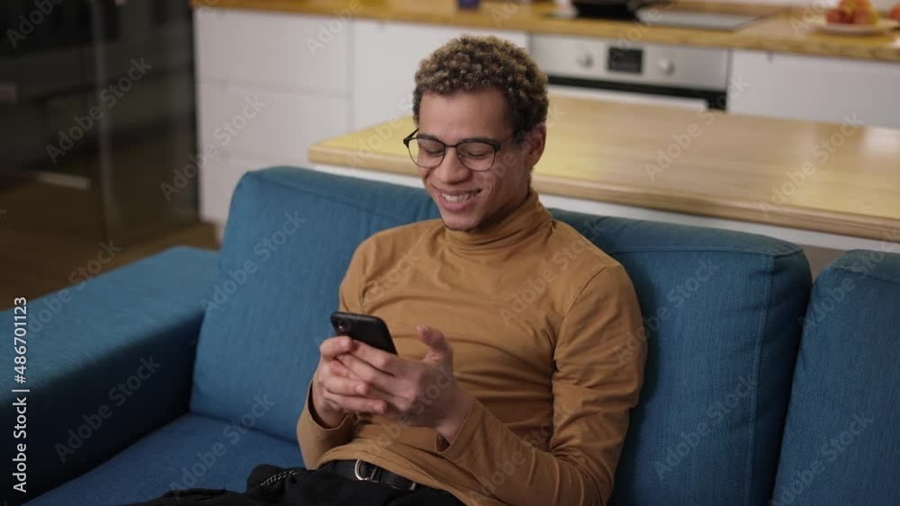 Curly young man web surfing on his mobile phone on the sofa at home Curly young man web surfing on his mobile phone on the sofa at home