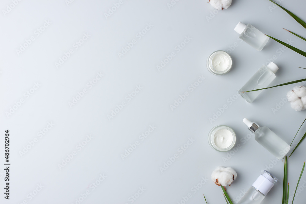 Fototapeta premium Cosmetic skin care products with flowers and palm leaf on grey background. Flat lay, copy space