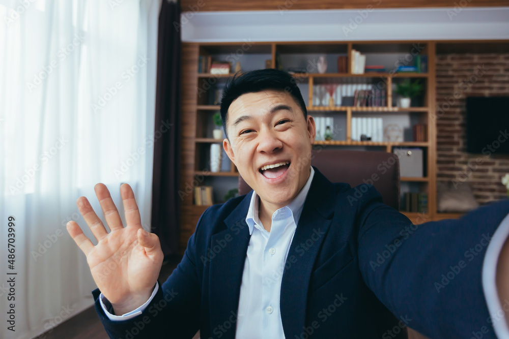 Cheerful and funny Asian businessman looks at the camera of a smartphone, talks on a video call, a man works in the office, waves a congratulatory gesture Cheerful and funny Asian businessman looks at the camera of a smartphone, talks on a video call, a man works in the office, waves a congratulatory gesture