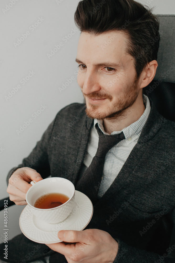a young stylish man with a beard and glasses in a shirt and a gray cardigan against a gray wall drinking coffee a young stylish man with a beard and glasses in a shirt and a gray cardigan against a gray wall drinking coffee