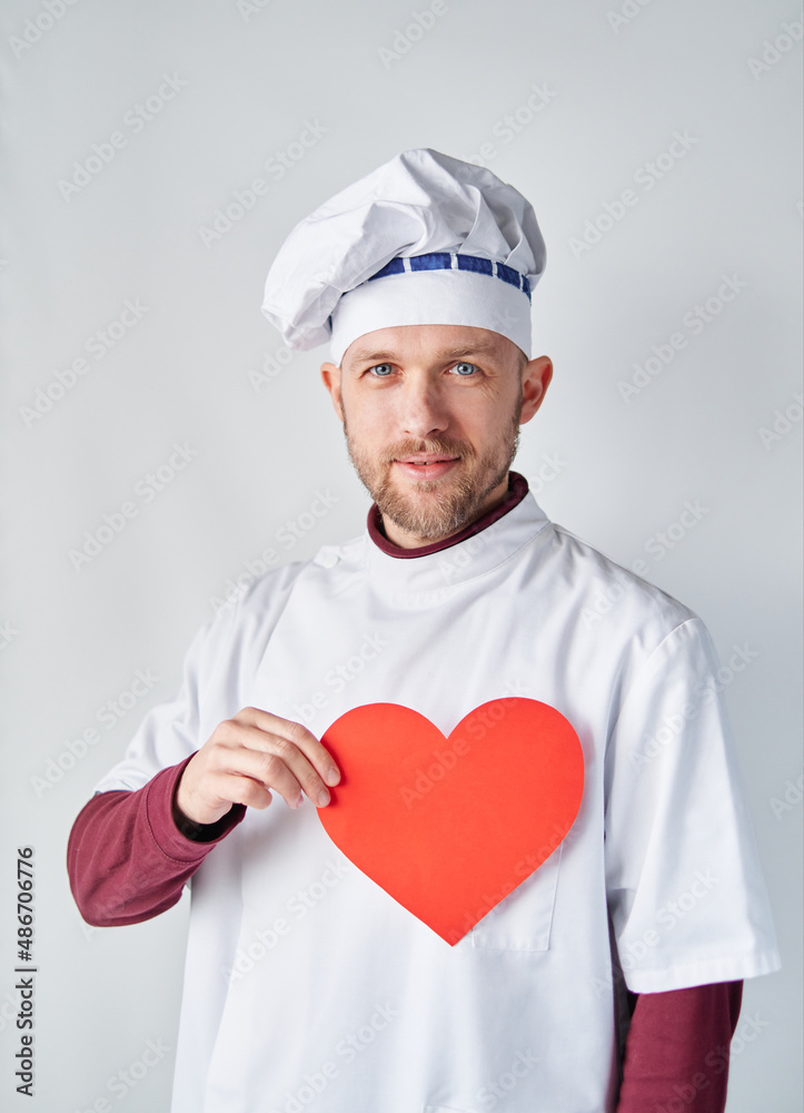Cheerful bearded male baker or chef in white uniform with red paper heart. Restaurant of cafe concept. Chef cook holding a red heart in front of the workwear. High quality photo Cheerful bearded male baker or chef in white uniform with red paper heart. Restaurant of cafe concept. Chef cook holding a red heart in front of the workwear. High quality photo