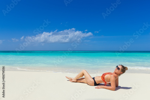 Fashion woman portrait in bikini on tropical beach. Relaxing summer mood, calm waves on sandy beach, exotic tropical island landscape with woman relaxing in the sand, sunglasses and sexy swimwear