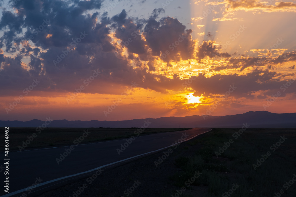 Beautiful sunset with cloudy sky and sun rays. Sunrise with road, mountains silhouettes and orange golden clouds.