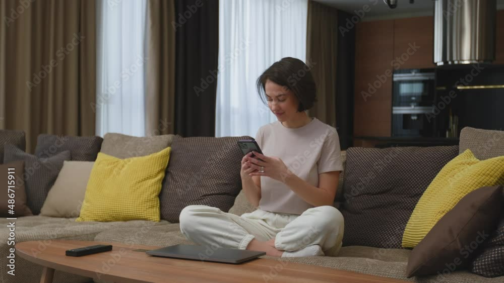 Happy young girl with phone using social network app chatting with friends or boyfriend sitting on sofa at home Happy young girl with phone using social network app chatting with friends or boyfriend sitting on sofa at home